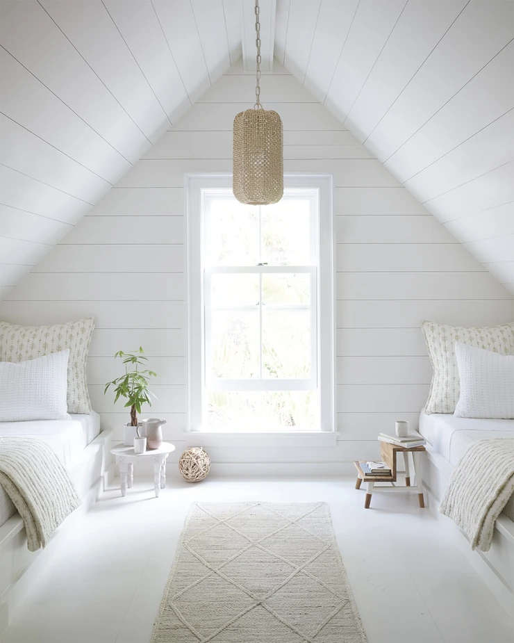 attic bedroom with natural light shiplap walls and vaulted a-frame ceiling two twin beds with wool rug runner natural beige sheets and pillow cases small wood side tables rattan hanging pendant light