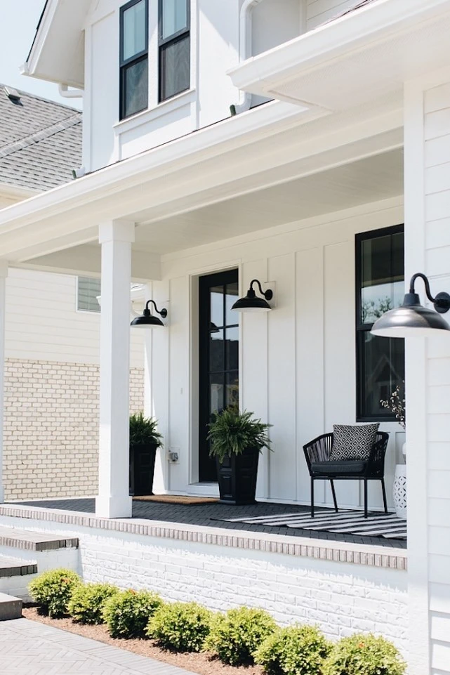 white board and batten exterior with wrap around porch black windows black gooseneck exterior lights wicker chair with black and white rug pots with plants at doorway boxwood in flower bed