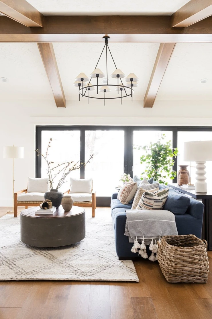 vaulted ceiling with wood details black framed accordion patio doors natural light wool area rug oversized blue sofa with throw pillows and throw basket on floor white table lamp floor lamp with two sitting chairs concrete grey ottoman oversized chandelier