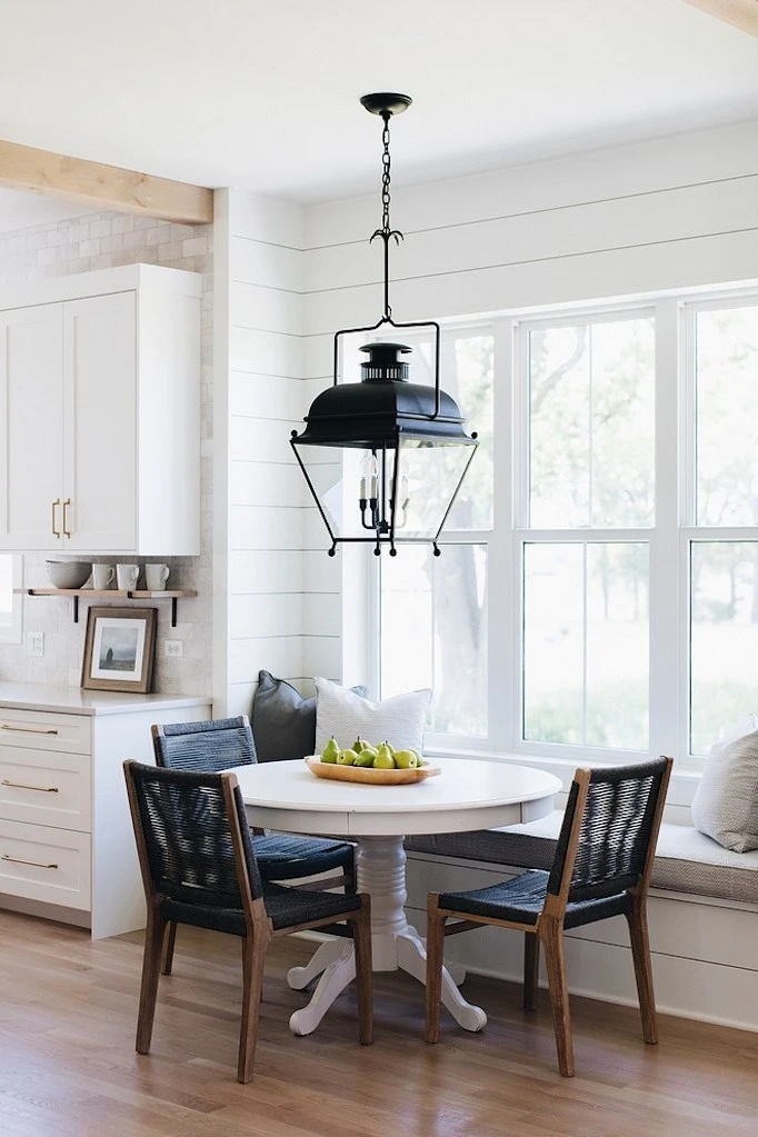 Kitchen nook with shiplap detail banquet seating round white table with three dark wood chairs with blue woven seats large black lantern pendant natural light windows kitchen eating area wood floors